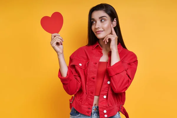 Thoughtful Young Woman Looking Paper Heart While Standing Yellow Background — Fotografia de Stock