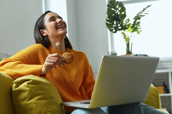 Happy Young Woman Using Laptop Enjoying Cookies While Sitting Couch —  Fotos de Stock