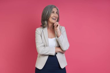 Senior woman in formalwear holding hand on chin and smiling while standing against pink background