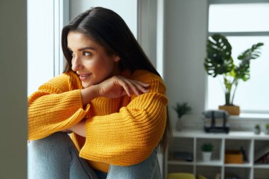 Dreamful young woman looking through a window and smiling while sitting on the window sill