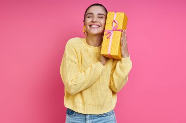 Happy young woman holding gift box near face and smiling against colored background