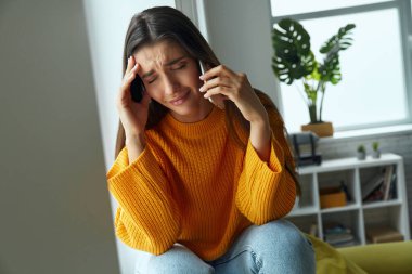 Depressed woman talking on mobile phone and touching head while sitting on the window sill