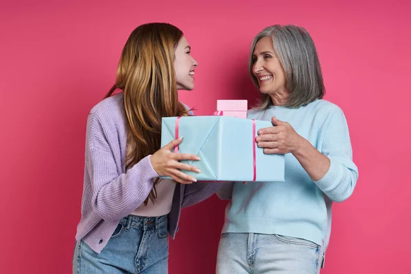 Happy senior mother receiving a gift box from her adult daughter against pink background
