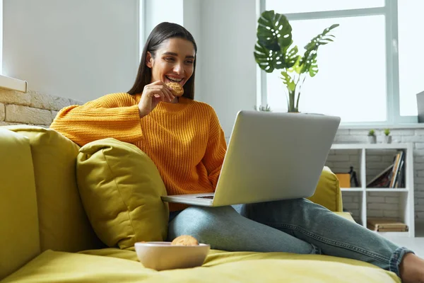 Attractive Young Woman Using Laptop Enjoying Cookies While Sitting Couch — стоковое фото