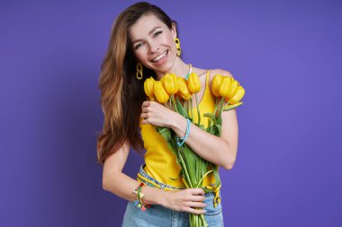 Beautiful young woman holding a bunch of tulips and smiling while standing against purple background