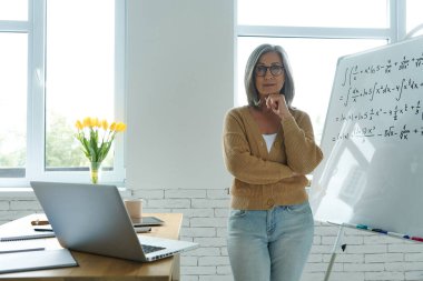 Confident senior woman holding hand on chin while standing near the whiteboard at classroom