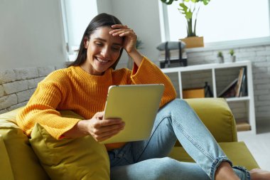 Beautiful young woman using digital tablet and smiling while sitting on the couch at home