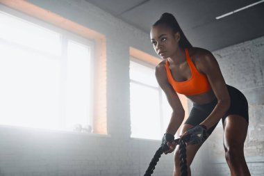 Concentrated young African woman preparing for training with battle ropes in gym