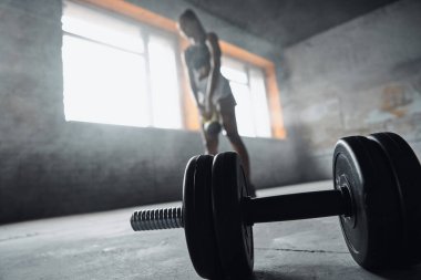 Concentrated young African woman exercising with kettlebell while dumbbell laying on foreground
