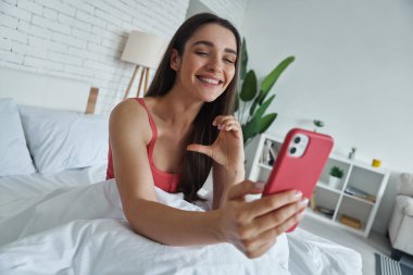 Beautiful young woman gesturing half heart while looking at smart phone and sitting in bed
