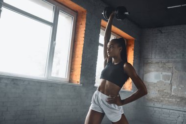 Confident young African woman using dumbbell while exercising in gym