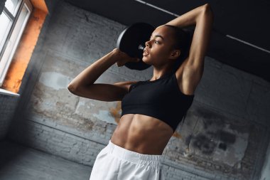 Confident young African woman working with weights while exercising in gym