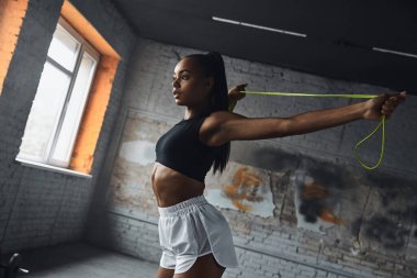 Confident young African woman using jumping rope while doing stretching exercises in gym