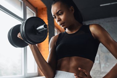 Concentrated young African woman using dumbbell while exercising in gym