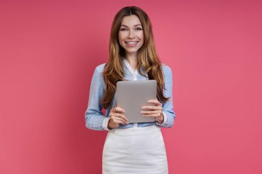 Confident young woman holding digital tablet and smiling while standing against colored background
