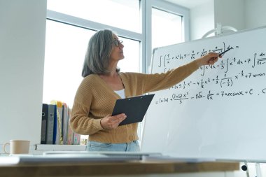 Confident senior woman teaching mathematics while pointing whiteboard at classroom