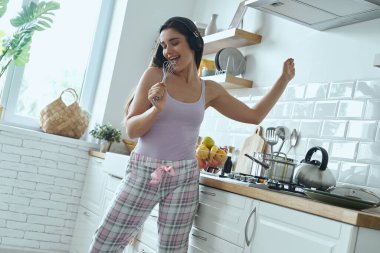 Happy young woman using whisk as microphone and dancing while cooking at the kitchen