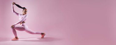 Attractive young woman in sports clothing doing stretching exercises against pink background