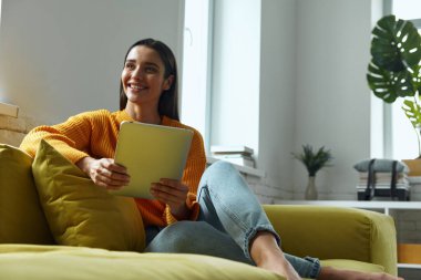 Attractive young woman using digital tablet and smiling while sitting on the couch at home