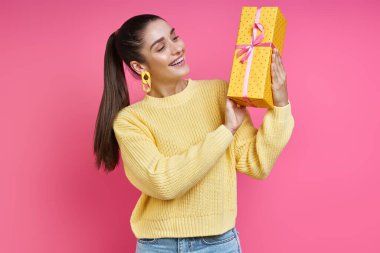 Happy young woman looking at gift box while standing against colored background