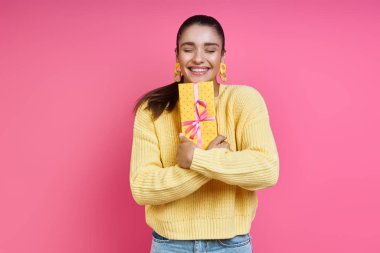 Excited young woman hugging gift box and smiling against colored background