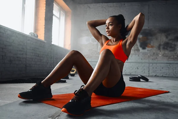 Beautiful young African woman adjusting her hair while sitting on exercise mat in gym