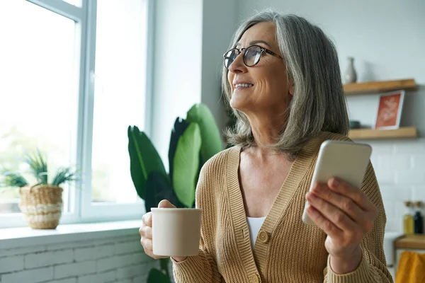 Cheerful Senior Woman Enjoying Hot Drink Holding Smart Phone While — Foto de Stock