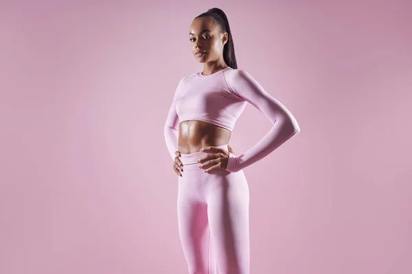 Confident young African woman in sports clothing standing against pink background