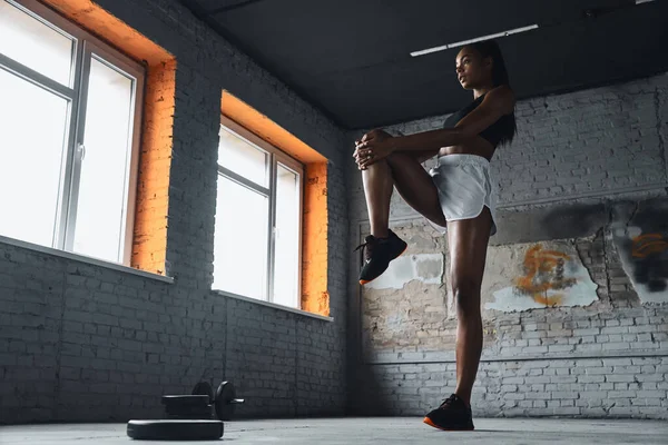 Full length of young African woman in sports clothing warming up before training in gym