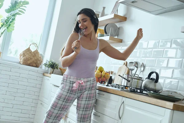 Happy young woman using whisk as microphone and dancing while cooking at the kitchen