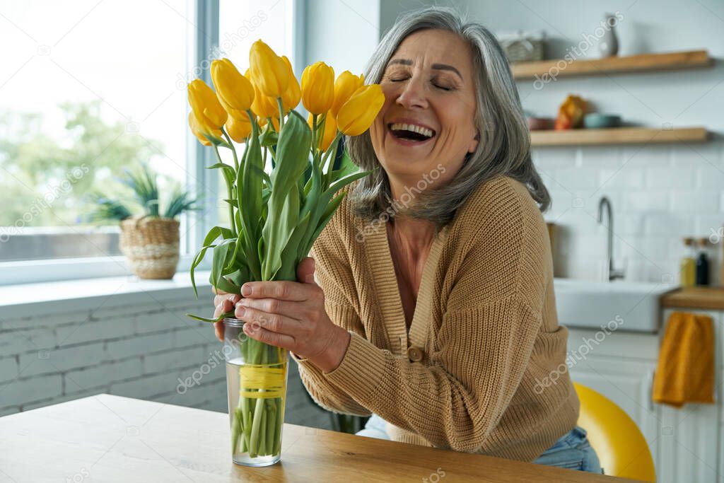 Senior woman holding a bunch of yellow tulips and looking happy while sitting at the kitchen island