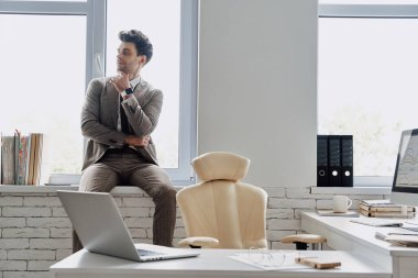 Thoughtful man in formalwear holding hand on chin while sitting on the window sill in office