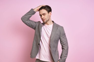 Handsome young man in suit holding hand in hair while standing against pink background
