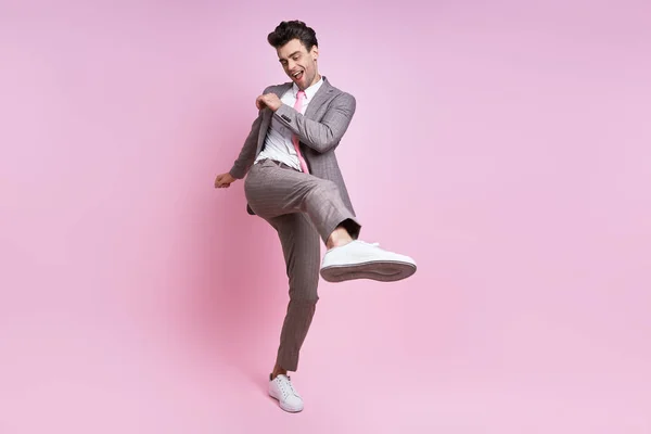 Happy young man in full suit throwing leg kick while standing against pink background