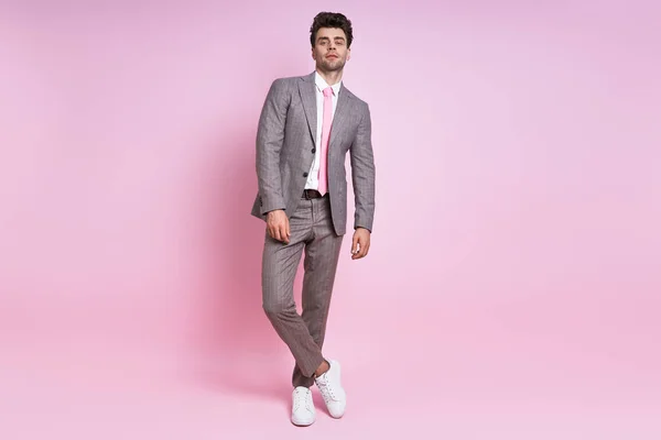 Confident young man in full suit looking at camera while standing against pink background