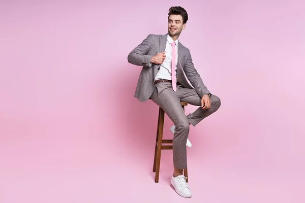 Handsome young man in full suit adjusting his jacket while sitting on chair against pink background