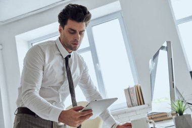 Confident young man in shirt and tie using technologies while working in office