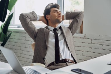 Relaxed young man in formalwear holding hands behind head while sitting in office