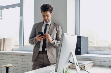 Confident young man using smart phone while standing near the window in office