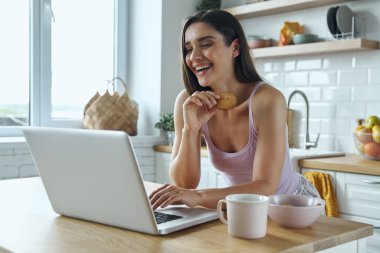 Happy young woman using laptop and eating cookies while sitting at the kitchen counter