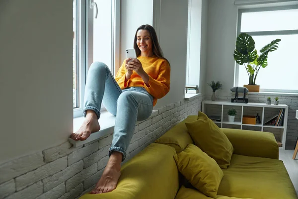 Happy Young Woman Using Smart Phone While Sitting Window Sill — Stok fotoğraf