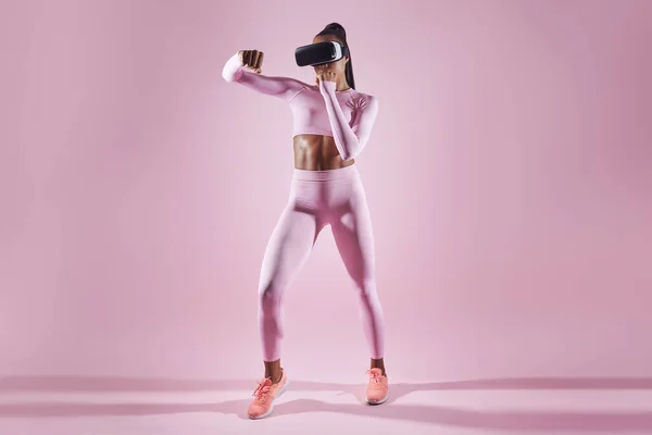 Woman in sports clothing wearing virtual reality headset while exercising against pink background