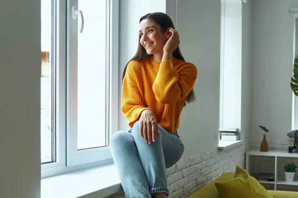 Happy young woman looking through a window while sitting on the window sill at home