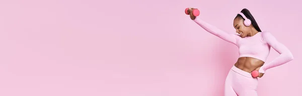 Happy African woman in headphones exercising with dumbbells against pink background