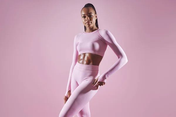 Attractive young African woman in sports clothing standing against pink background