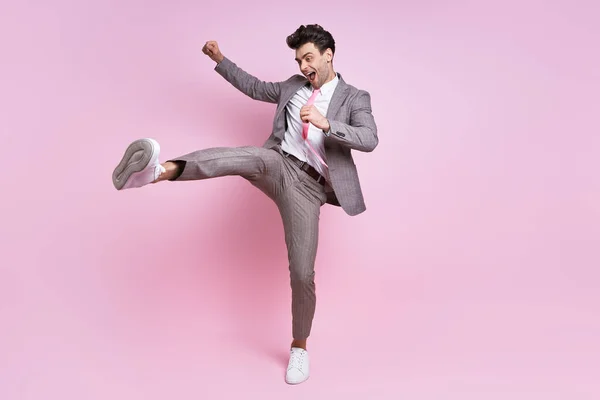Excited young man in full suit throwing leg kick while standing against pink background
