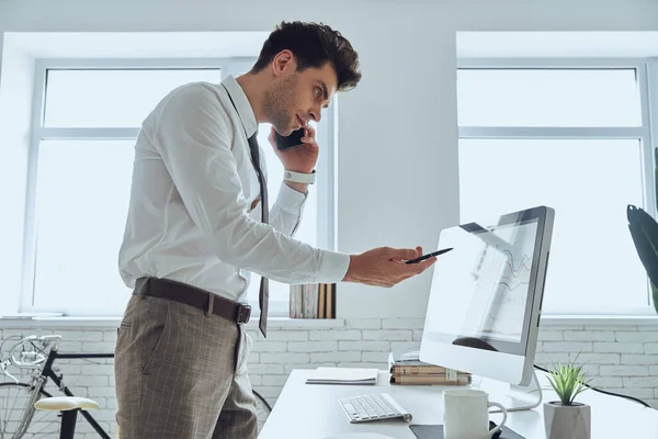 Concentrated man looking at the computer monitor and pointing it while talking on phone in office