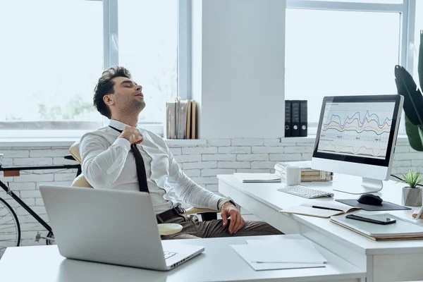 Tired Young Man Adjusting His Necktie While Sitting His Working — Stockfoto