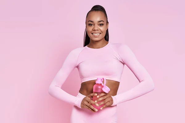 Happy African Woman Sports Clothing Holding Bottle Water Pink Background — Stockfoto