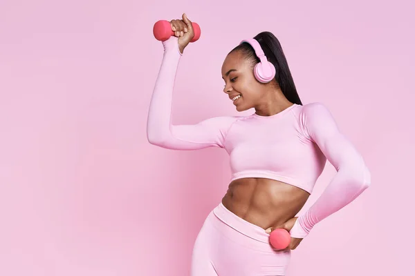 Beautiful African woman in headphones exercising with dumbbells against pink background